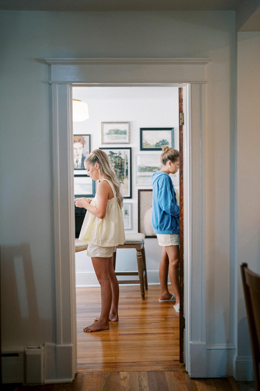 Two young women standing back-to-back in a brightly lit room with wooden floors, wearing casual summer clothing. One is in a yellow top near a wall adorned with artwork and photographs.
