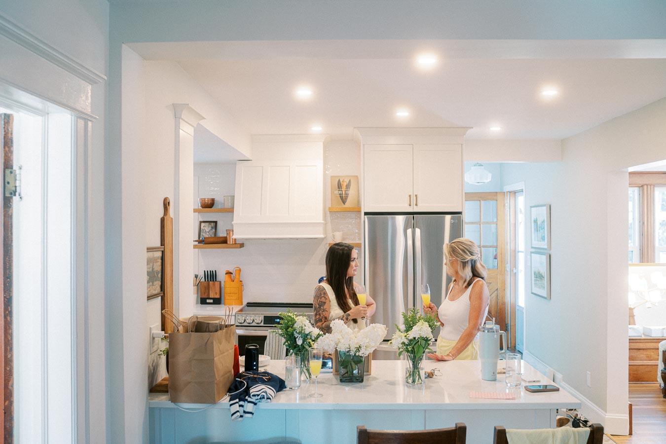 Two women conversing in a bright, modern kitchen with white cabinets, stainless steel appliances, and a floral centerpiece.