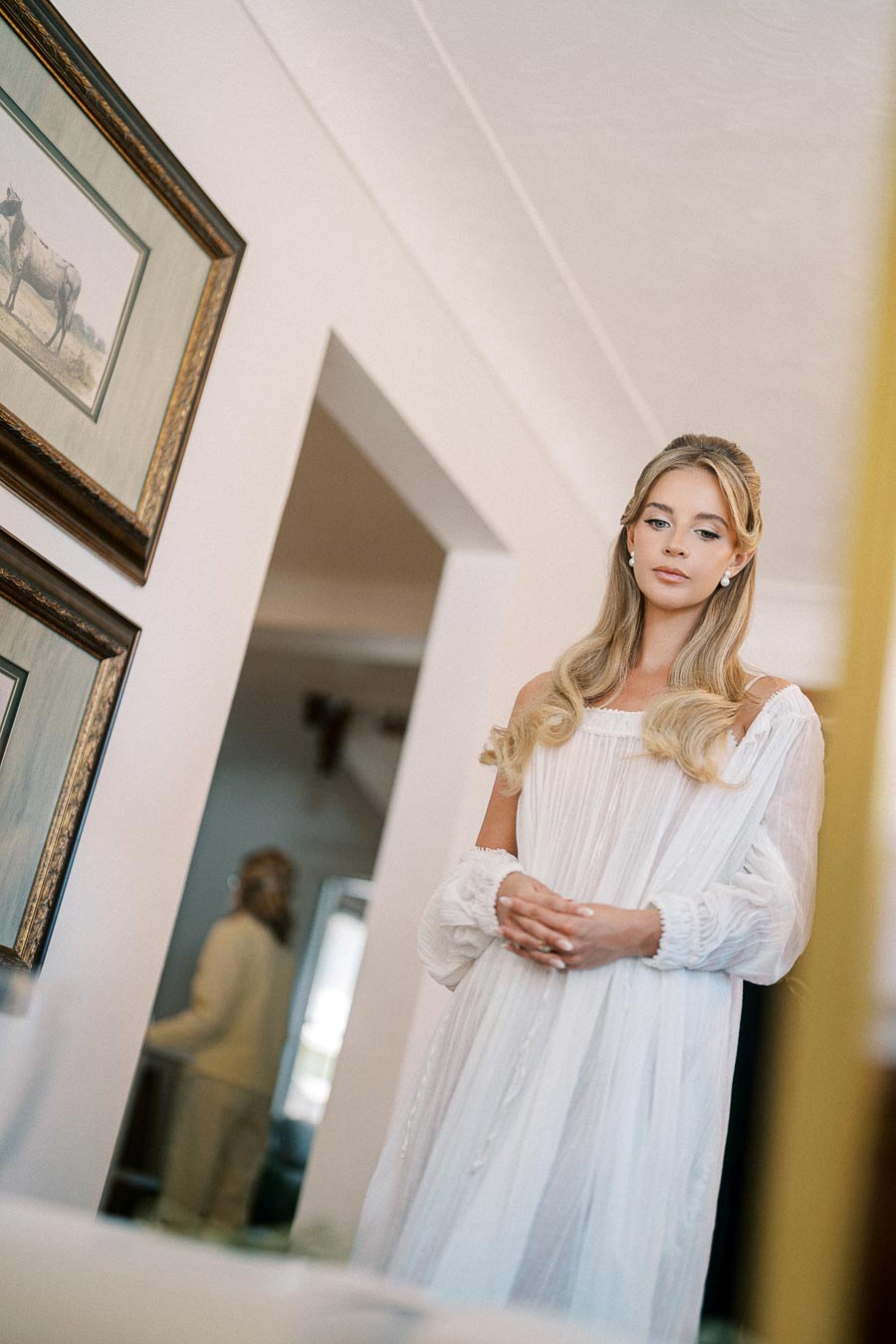 Elegant woman in a flowing white dress standing in a well-lit room with framed art on the walls.