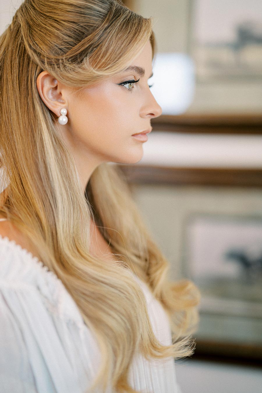 A woman with long blonde hair styled elegantly, wearing pearl earrings and a white blouse, looks thoughtfully to the side.