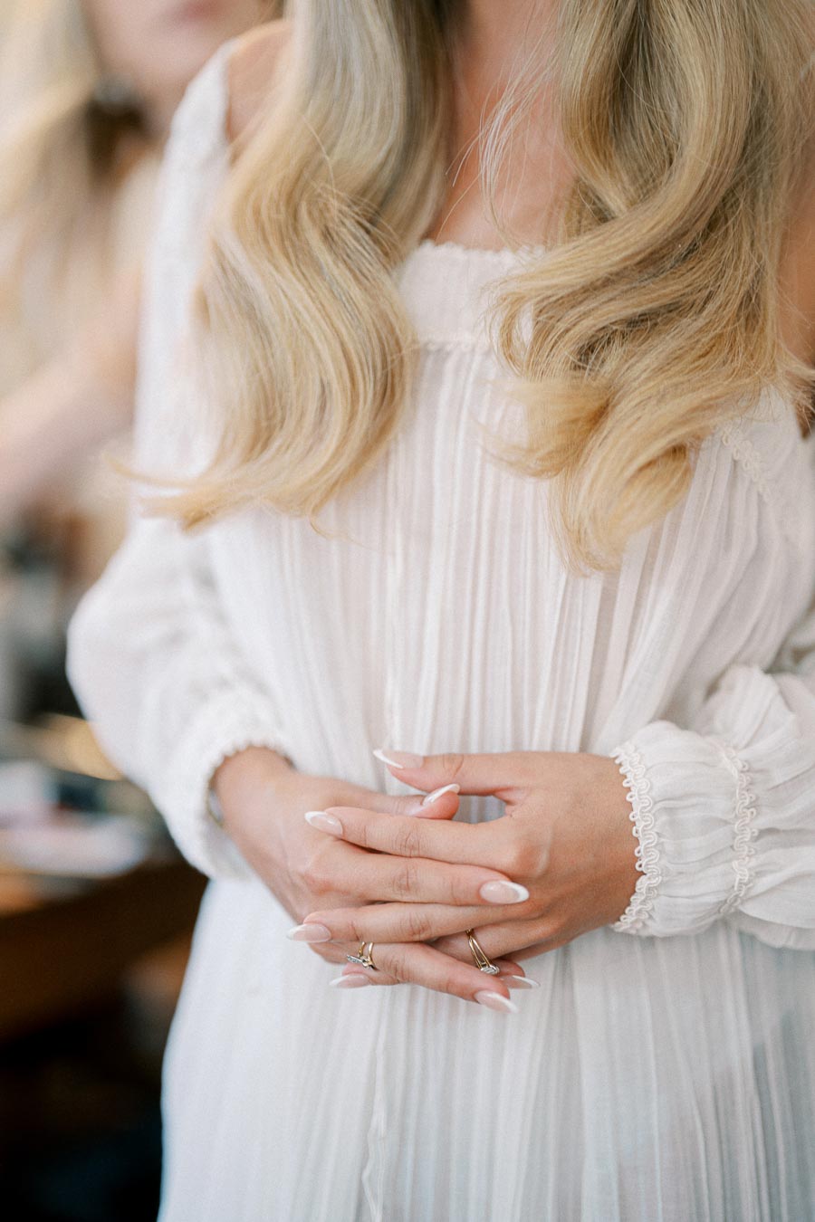 Elegant woman in a flowing white dress with long, wavy blonde hair and manicured nails, crossing her hands displaying simple rings.