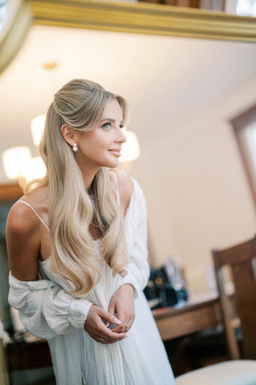 A bride with long, wavy blonde hair and pearl earrings gazing into a mirror, wearing an elegant wedding gown in a softly lit room.