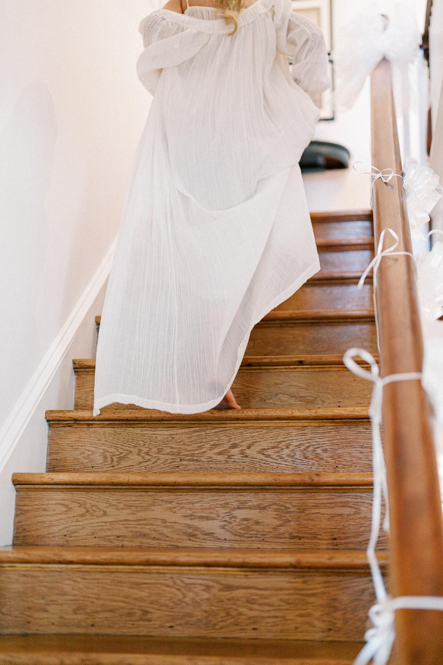 A person in a flowing white dress walking up a wooden staircase adorned with delicate white ribbons, creating an elegant and serene atmosphere.