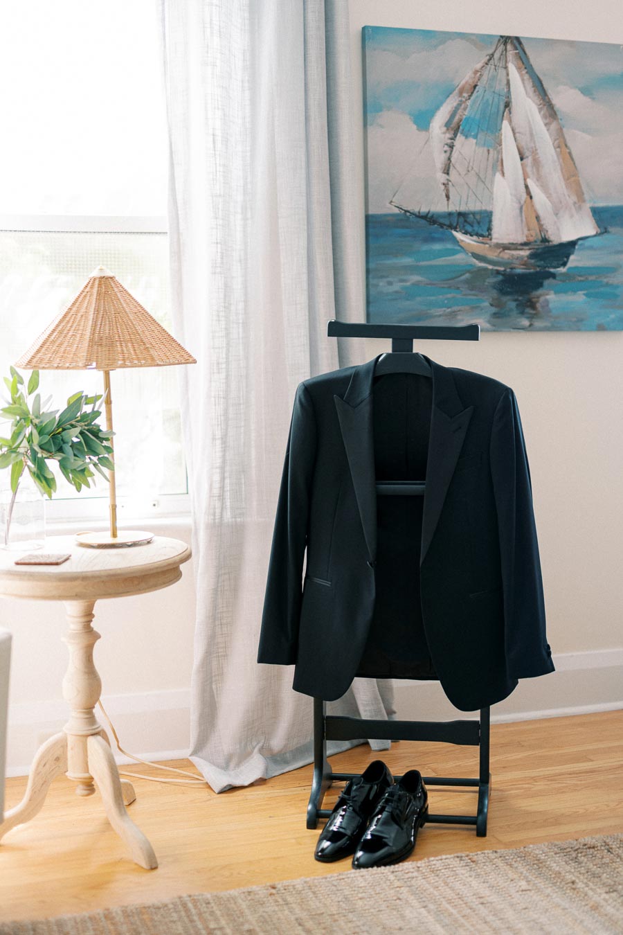 Formal black tuxedo jacket and polished shoes on a valet stand, next to a wooden side table with a woven lamp, with a nautical painting in the background.