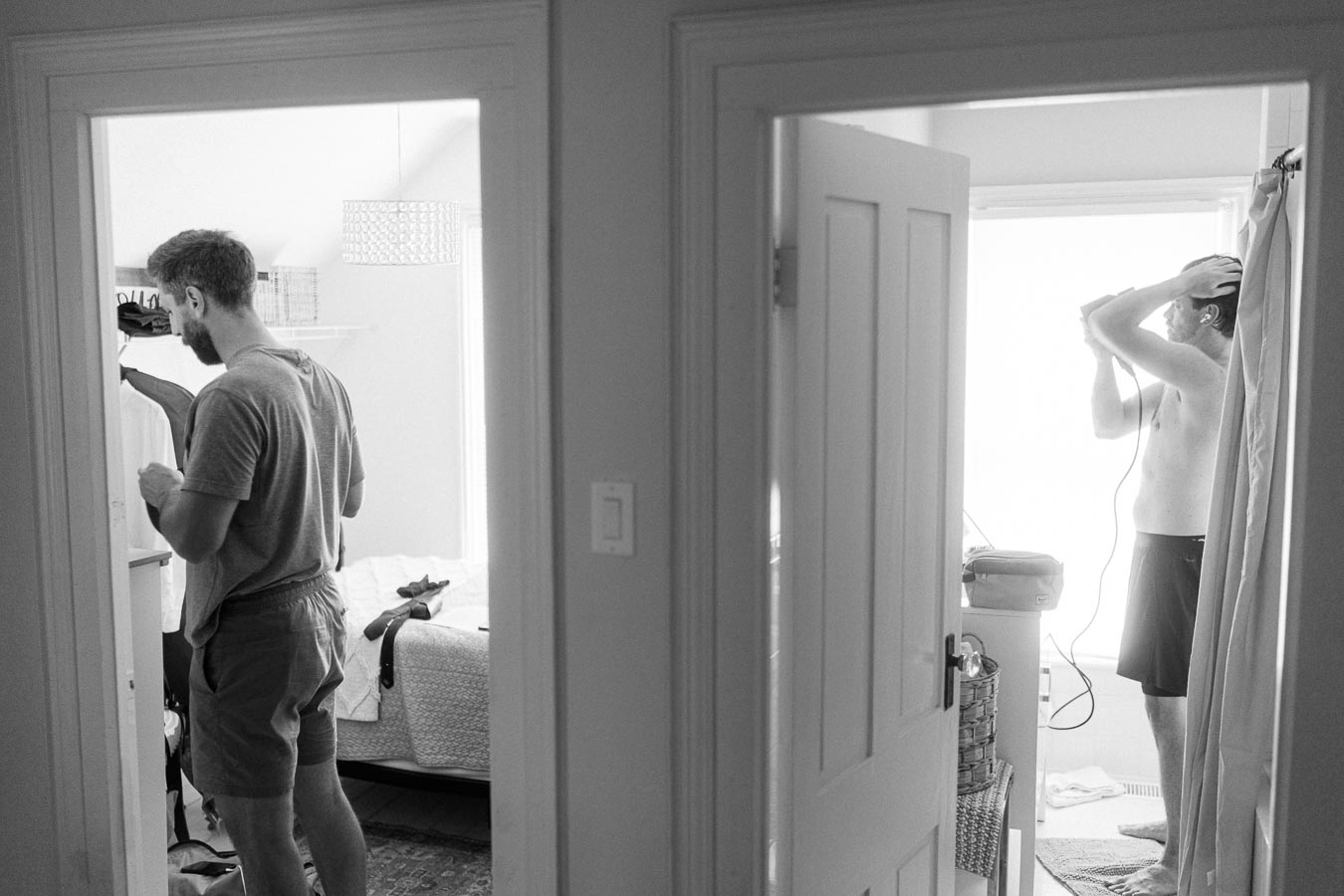 Black and white photo of two men in adjacent rooms of a house. One is drying his hair in a bathroom, while the other selects clothes in a bedroom. The scene captures a candid morning routine.
