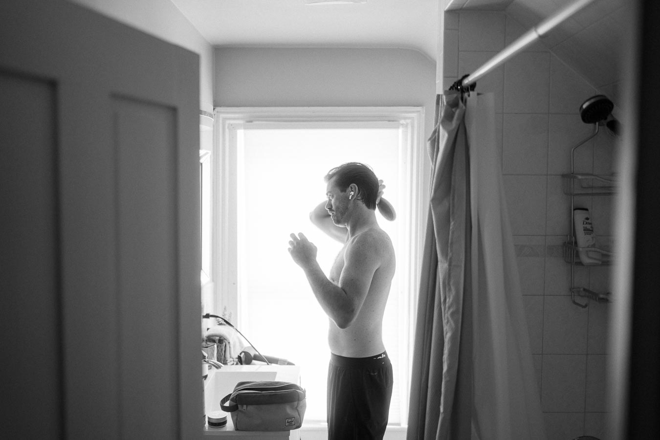 Black and white image of a shirtless man grooming his hair in a brightly lit bathroom, with a grooming kit on the sink and a shower area in the background.