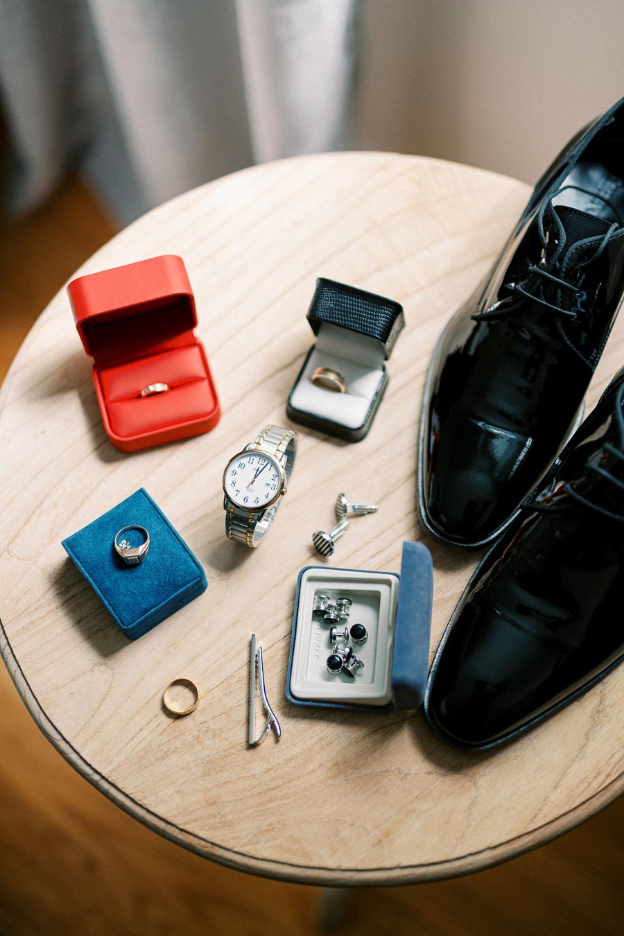 Elegant wedding preparation essentials on a wooden table, featuring shiny black dress shoes, a classic wristwatch, cufflinks, tweezers, and three ring boxes, creating a refined and sophisticated ensemble for a groom.