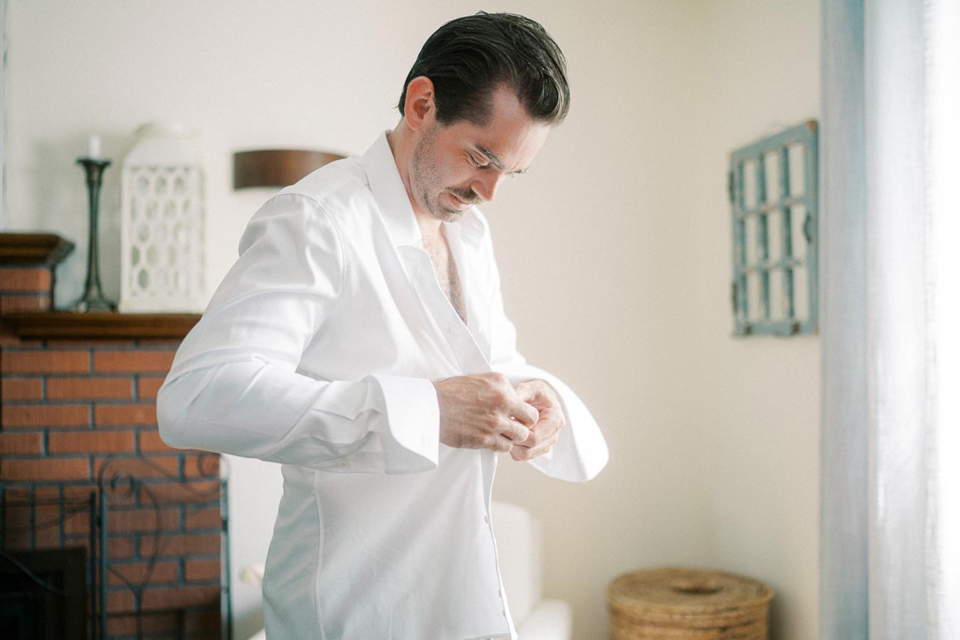 Man in a white shirt buttoning up in a softly lit room, showcasing a calm and focused demeanor near a brick fireplace.