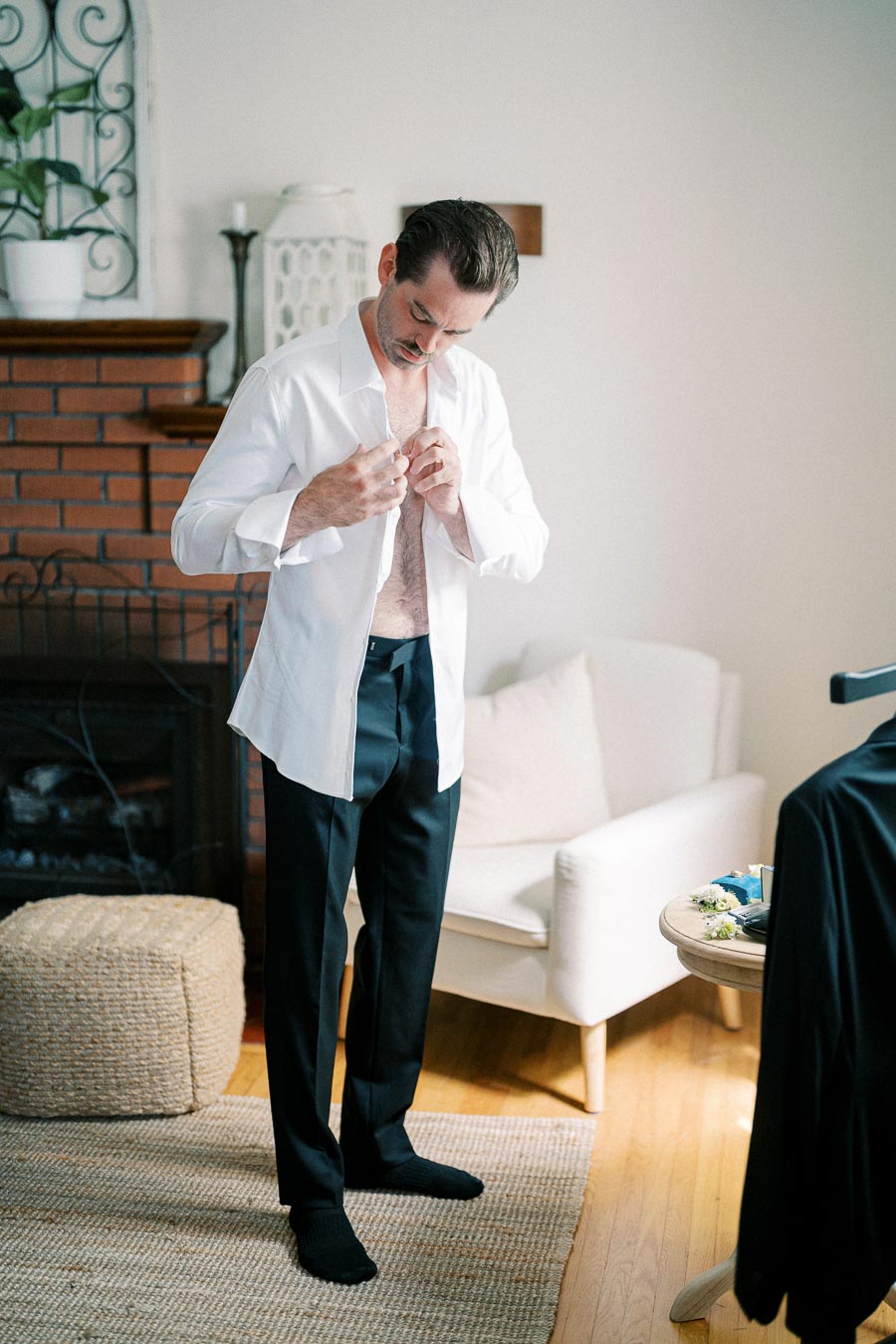 Man buttoning a white dress shirt in a cozy living room setting, preparing for a formal event.