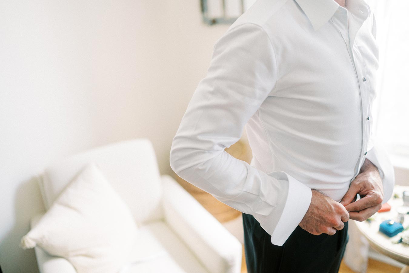 Man adjusting formal white shirt before an event; tailored attire preparation in bright interior setting with a white armchair.