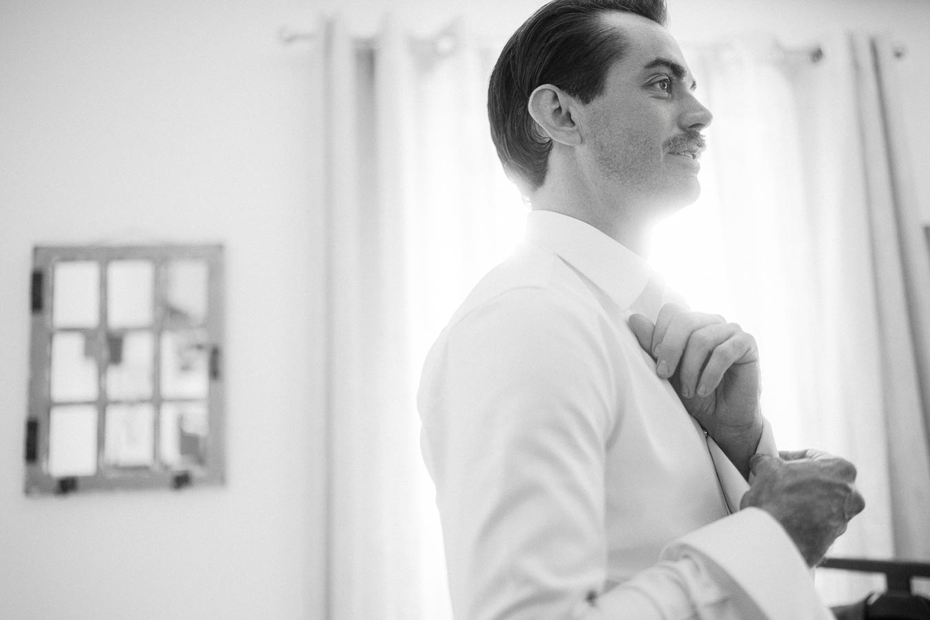Black and white image of a groom adjusting his tie in preparation for his wedding, with natural light streaming in from a nearby window creating a soft, elegant atmosphere.