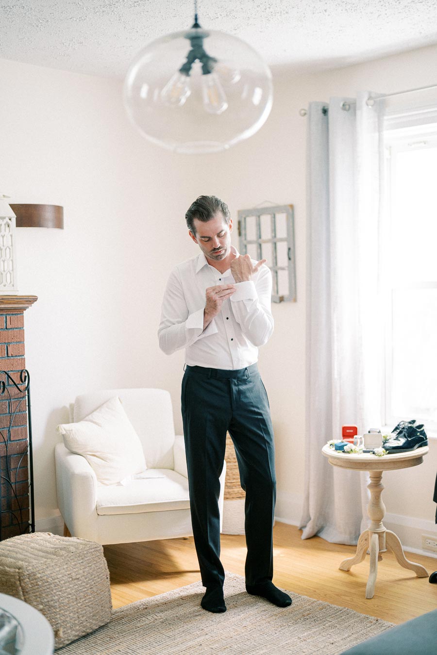 A man in a white dress shirt adjusts his cufflinks in a bright, stylish living room with a modern, cozy decor, featuring a white armchair, a round wooden table with accessories, and a vintage hanging light fixture.