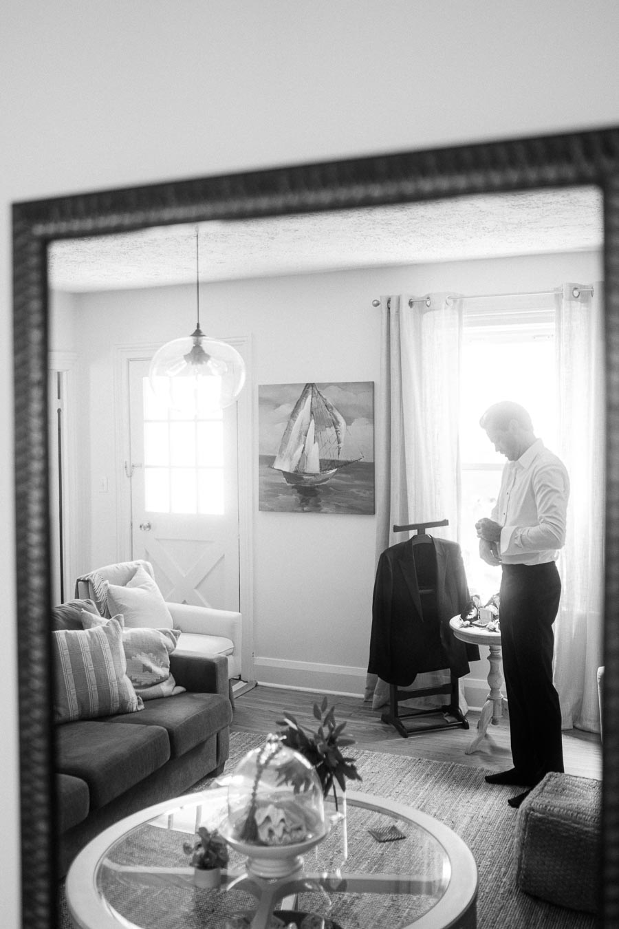 Black and white image of a stylish living room showing a man adjusting his cuffs in front of a window, with a suit jacket on a stand next to him. The room features a sailing ship painting, a modern pendant light, a comfortable sofa with patterned cushions, and a glass tabletop with decorative items, all reflected in a large mirror.