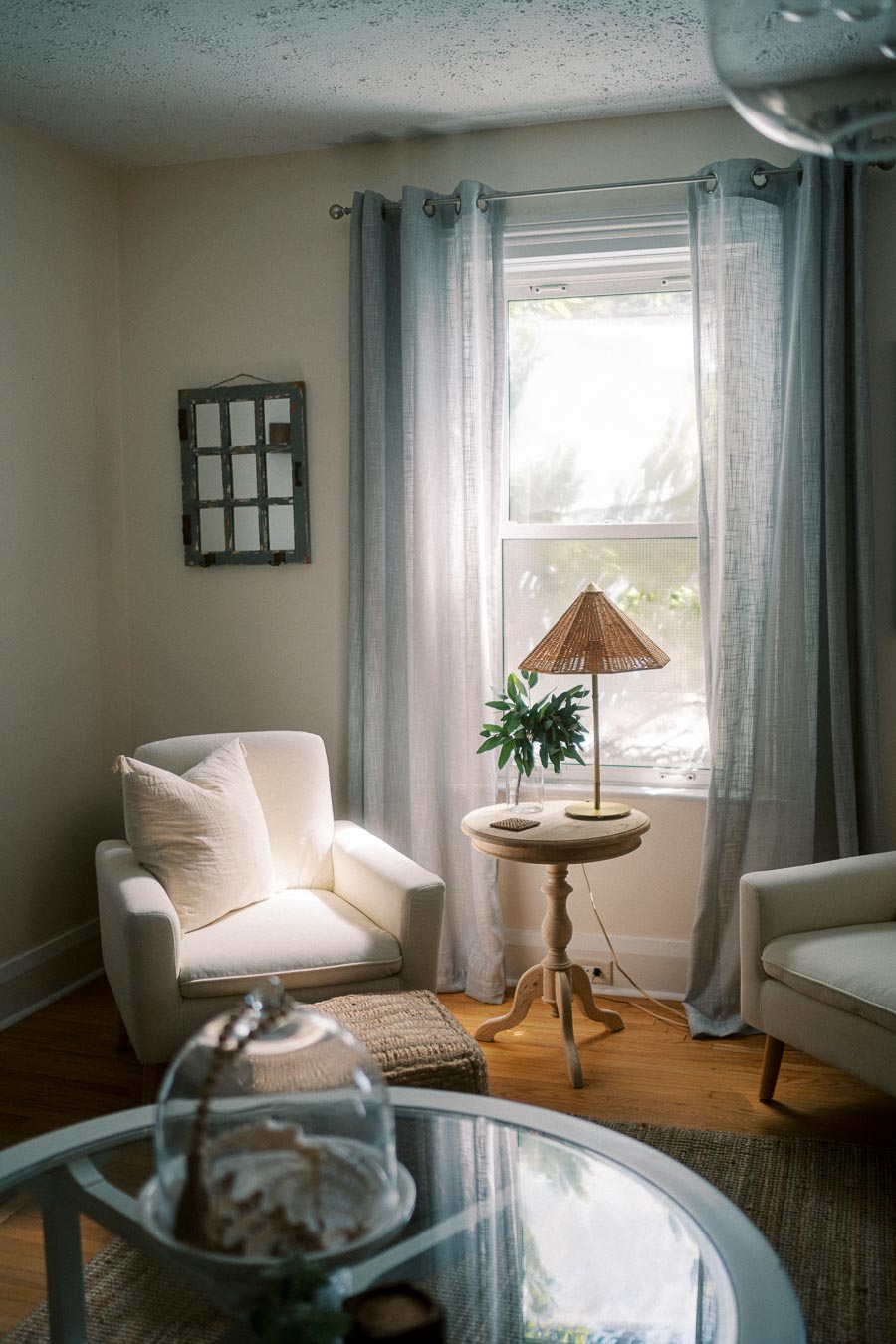 Cozy living room with natural light streaming through a window with sheer curtains, featuring a white armchair, a wicker lamp, a small plant on a round wooden table, and a decorative glass cloche on a coffee table.
