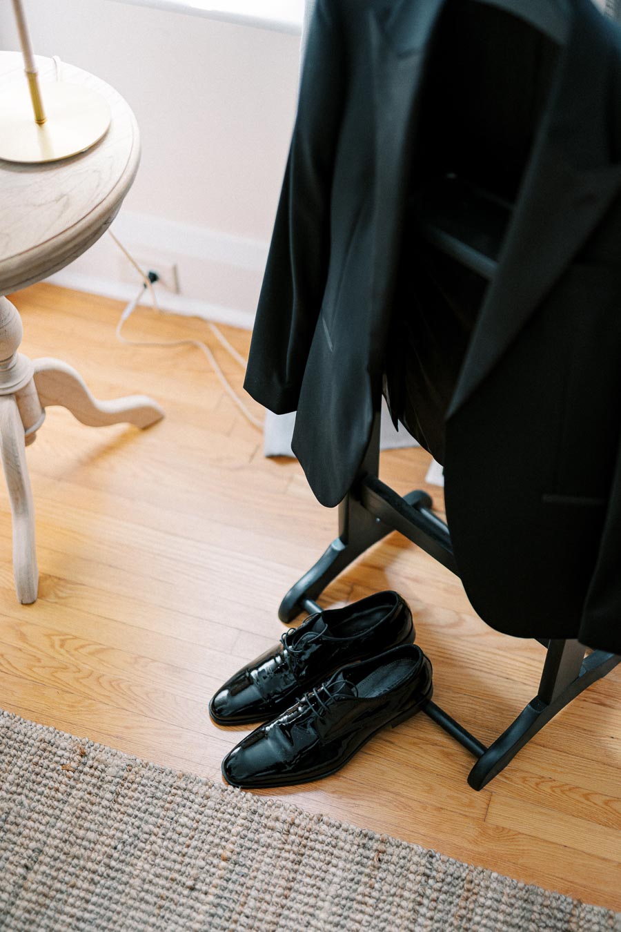 Black formal suit jacket on a chair with polished black dress shoes on a hardwood floor, next to a wooden table with a beige rug.