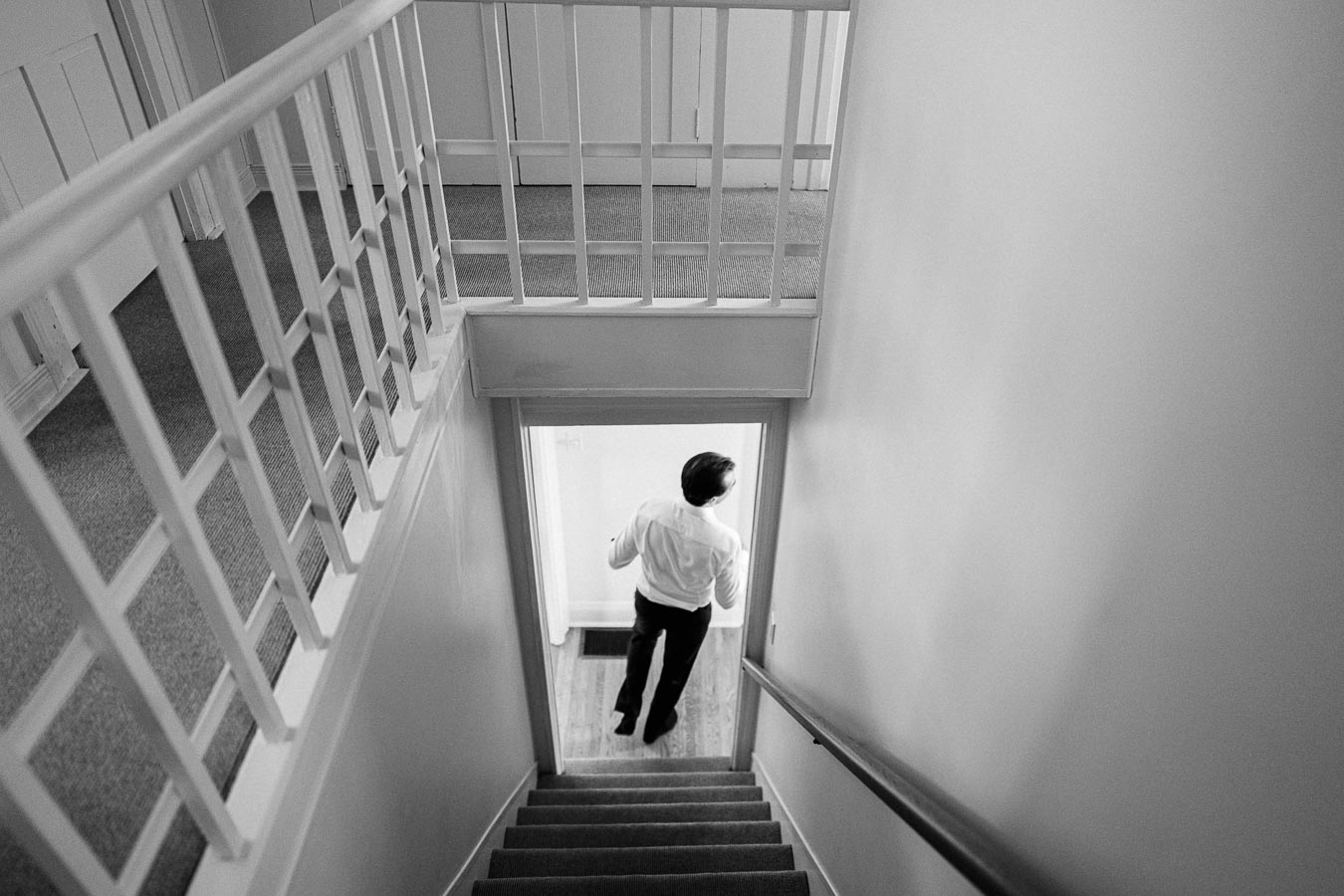 Black and white image of a person in formal attire descending a carpeted staircase, leading to a well-lit room.