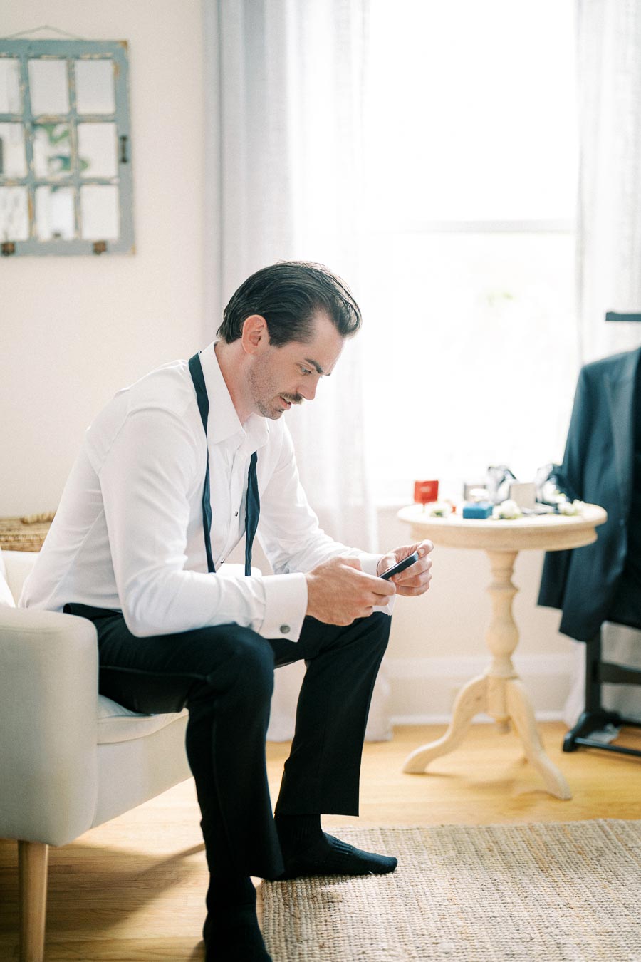 A man dressed in formal attire sits on a white chair, looking at his smartphone. He is wearing a white dress shirt with suspenders, black pants, and black socks. The room is well-lit, with a small table in the background holding various accessories and a jacket hanging on the back of a chair. Bright, natural light filters through a window