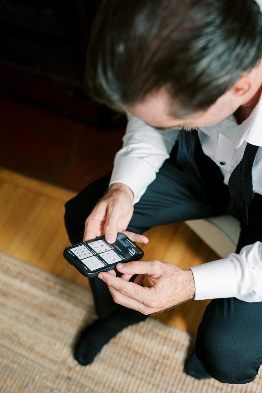 A man in formal attire checks his smartphone, engrossed in an app displaying multiple icon options, suggesting a focus on organization or technology in a home setting.