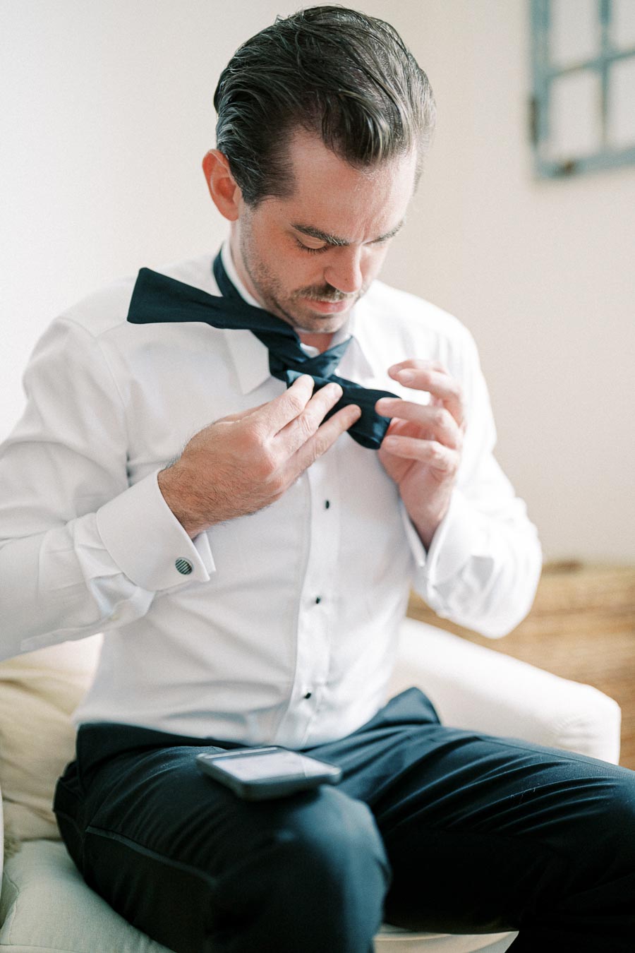 Man in a formal white shirt adjusting a black bow tie while seated, preparing for a formal event or wedding.