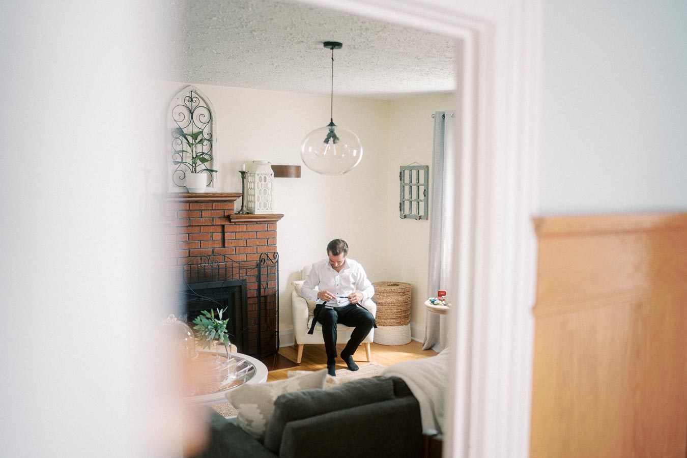 Man sitting in a cozy living room with a white armchair and a brick fireplace, reading a book near natural light from a window.