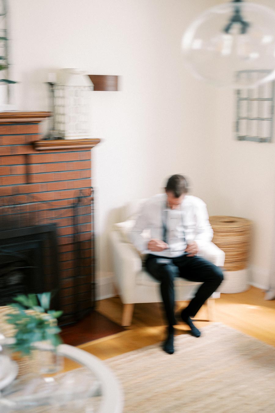 Blurry image of a person sitting in a cozy, well-lit living room with a red brick fireplace and a white cushioned chair, conveying a relaxed home environment.