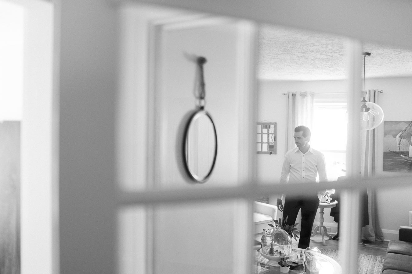 Black and white photo of a man in a white shirt standing in a living room, viewed through a window. The room features modern decor with a round table and a large picture window.