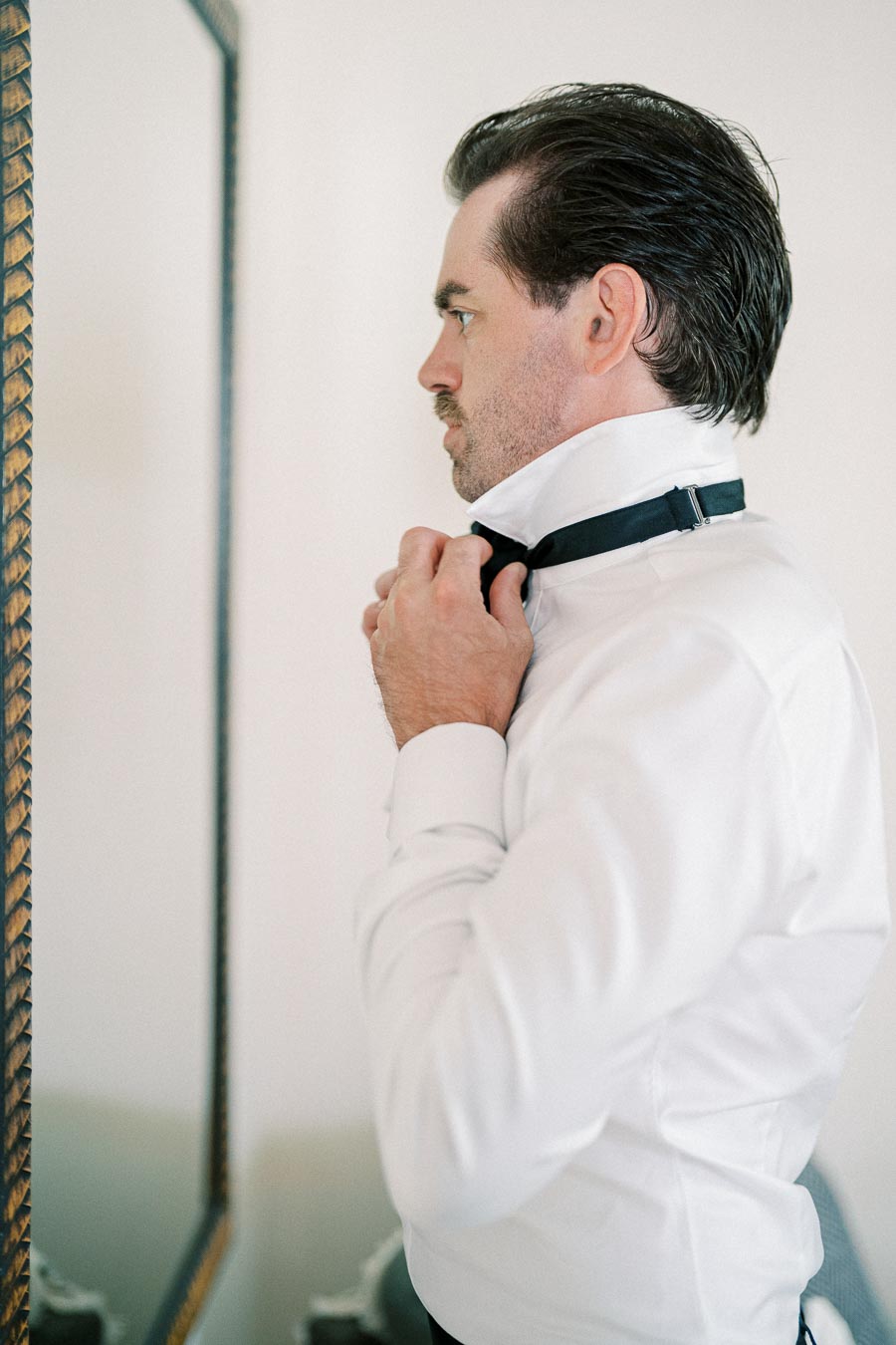 Man adjusting a black bow tie in front of a mirror, wearing a white dress shirt.