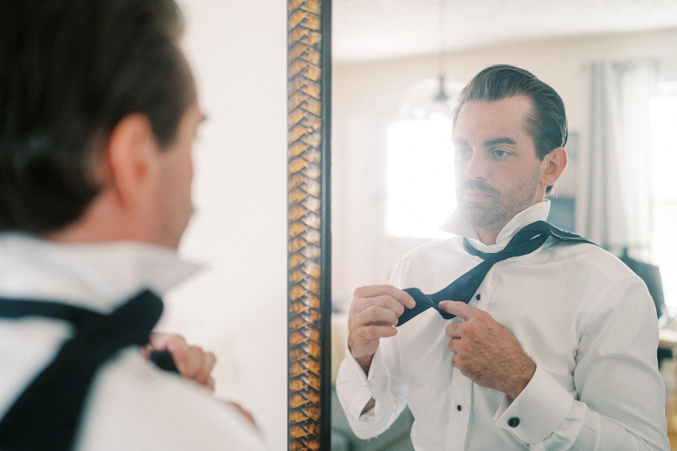 Man in a white dress shirt tying a black tie in front of a mirror, preparing for a formal event.
