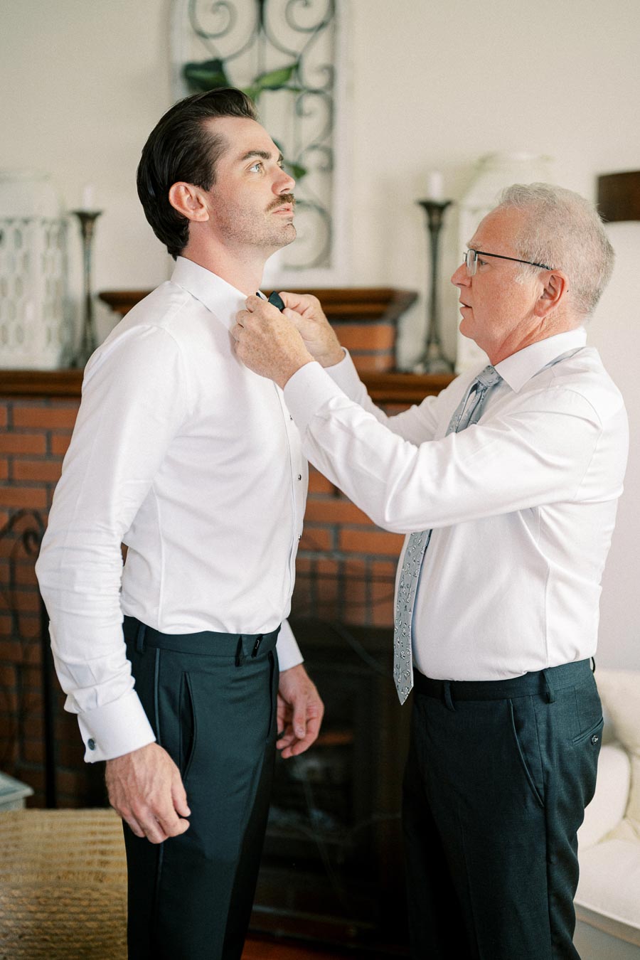 A groom getting his bow tie adjusted by an older gentleman before a wedding ceremony in a warmly decorated room.