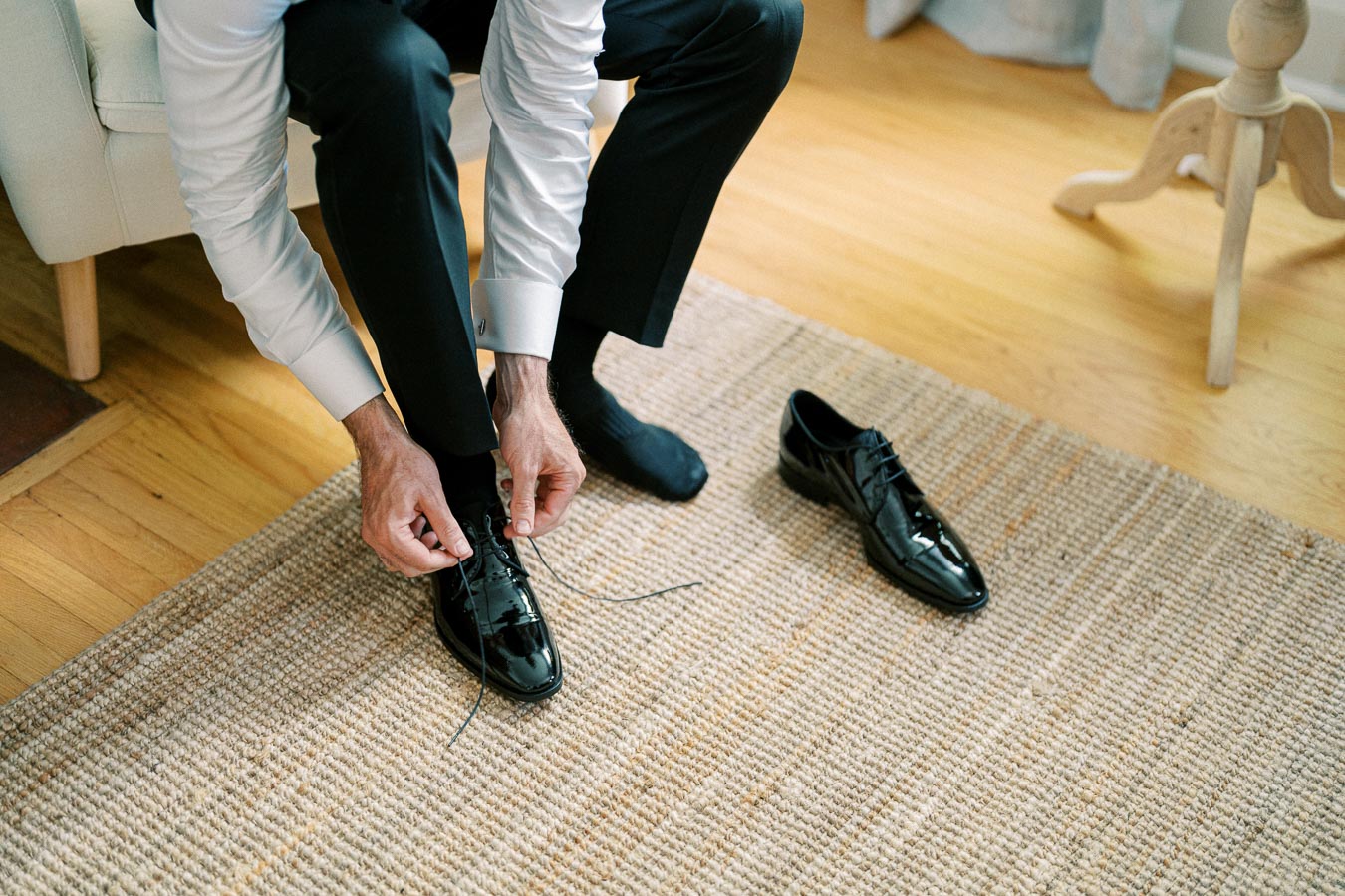 Man in formal attire tying black dress shoes on a woven rug, preparing for a formal event or wedding.