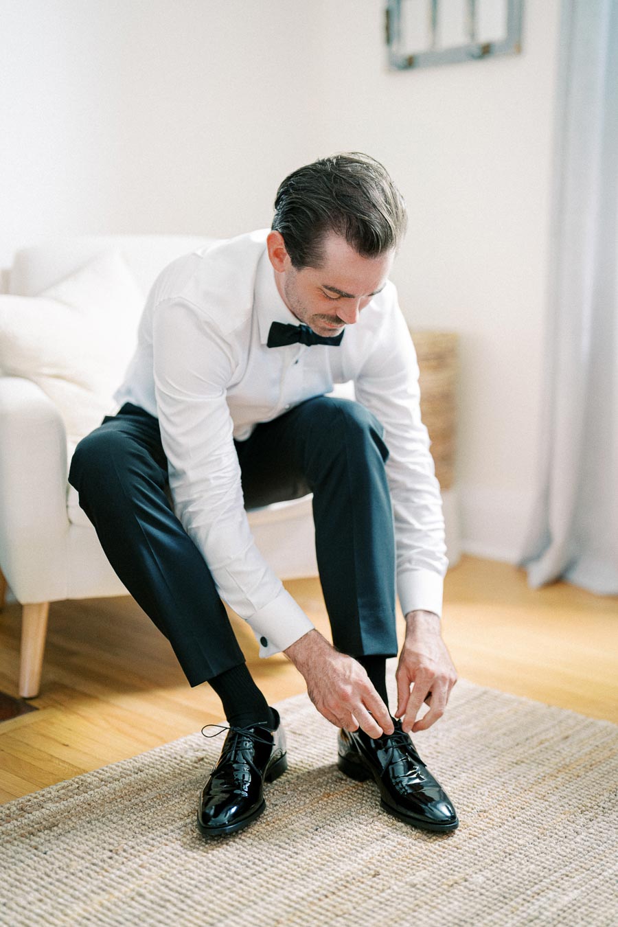 A groom in a formal white shirt and black bow tie, sitting on a chair and tying his black dress shoes in preparation for a wedding, in a bright room with natural light.