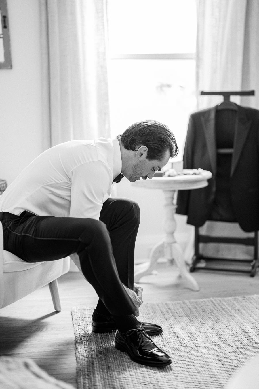 Black and white image of a man sitting on a chair, wearing formal attire, tying his shoe in a well-lit room with a suit jacket hanging in the background.