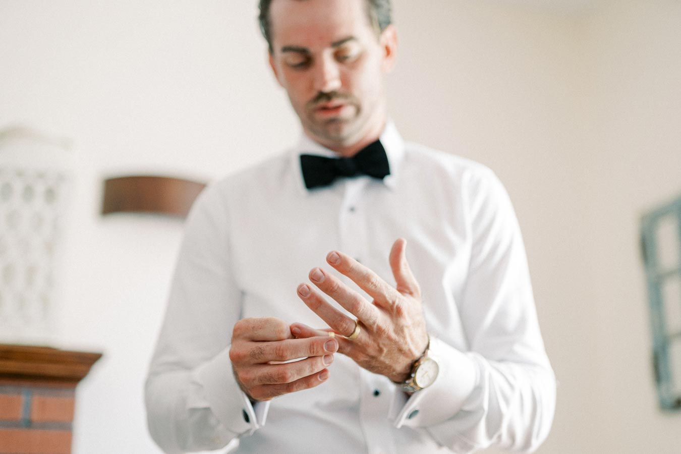 A man in a white dress shirt and black bow tie adjusting his wedding ring, standing indoors with soft lighting, focusing on his hands and elegant watch.