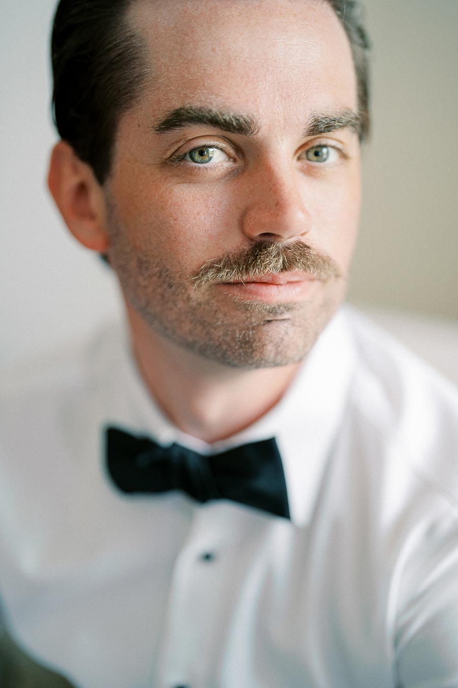 A close-up portrait of a person with a mustache, wearing a white dress shirt and black bow tie, looking relaxed and confident.