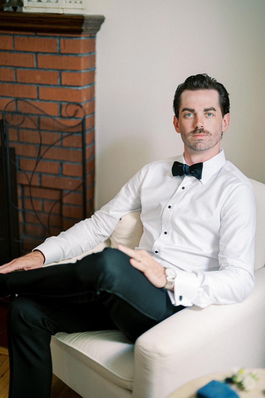 A man sitting in a white chair wearing a white dress shirt and black bow tie, with a brick fireplace in the background, exuding a formal and sophisticated look.