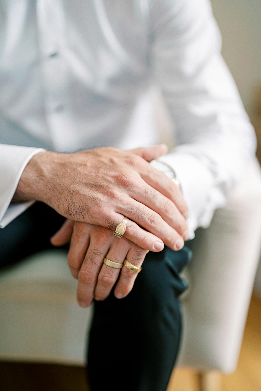 A man wearing a white dress shirt sits with hands clasped, showcasing multiple gold rings with intricate designs, highlighting modern men's jewelry.