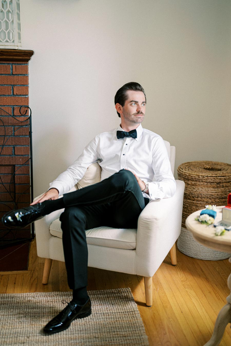 A groom sitting on a white chair indoors, wearing a formal white shirt and black bow tie, preparing for his wedding day.