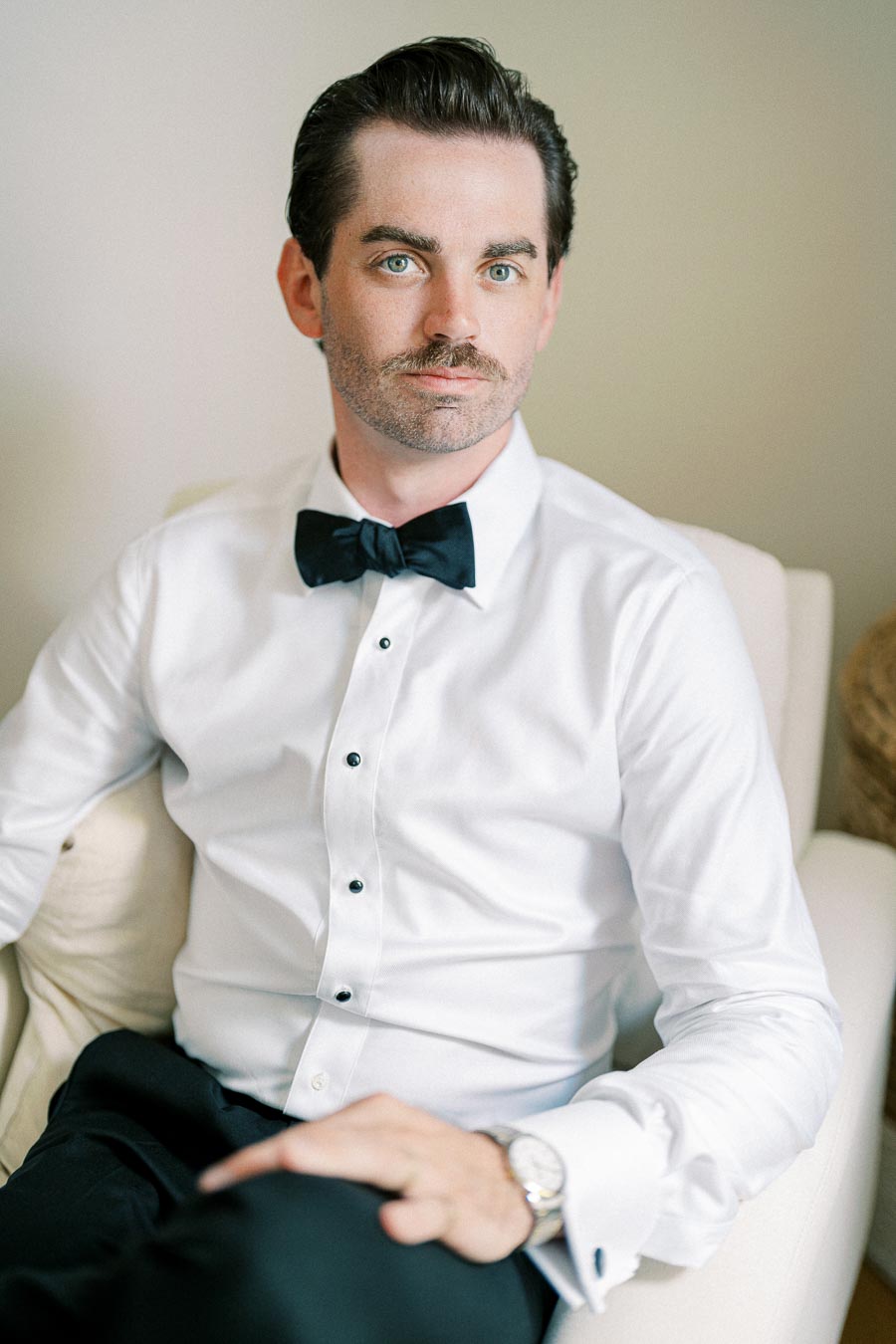 Elegant man in a white dress shirt and black bow tie sitting on a beige chair, showcasing formal attire and style.