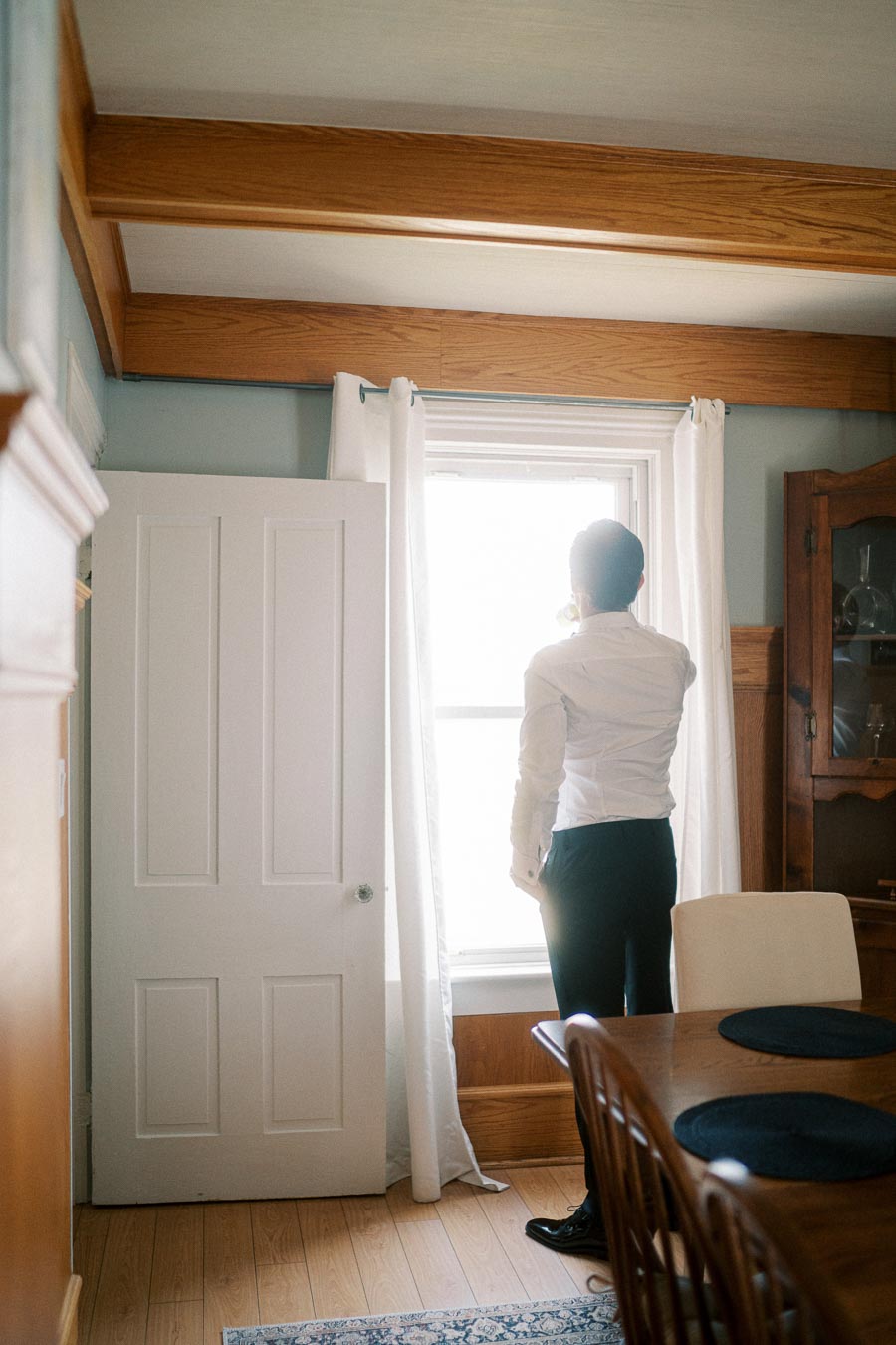 Man in formal attire standing by a window in a cozy dining room, daylight streaming through white curtains, with wooden furniture and blue accents.