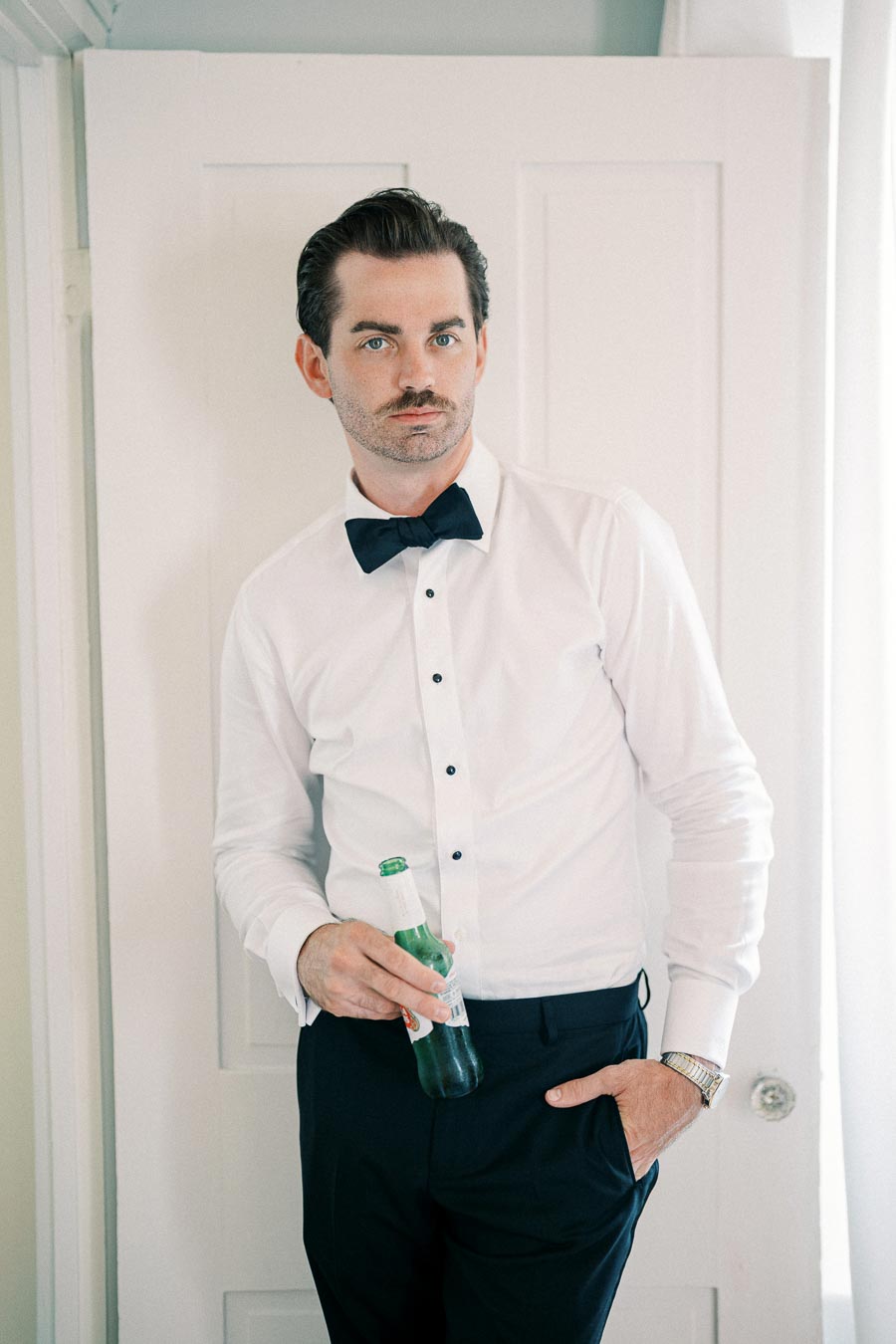 A man in formal attire with a white shirt, black bow tie, and black pants holds a green glass bottle, standing casually against a white wooden door.