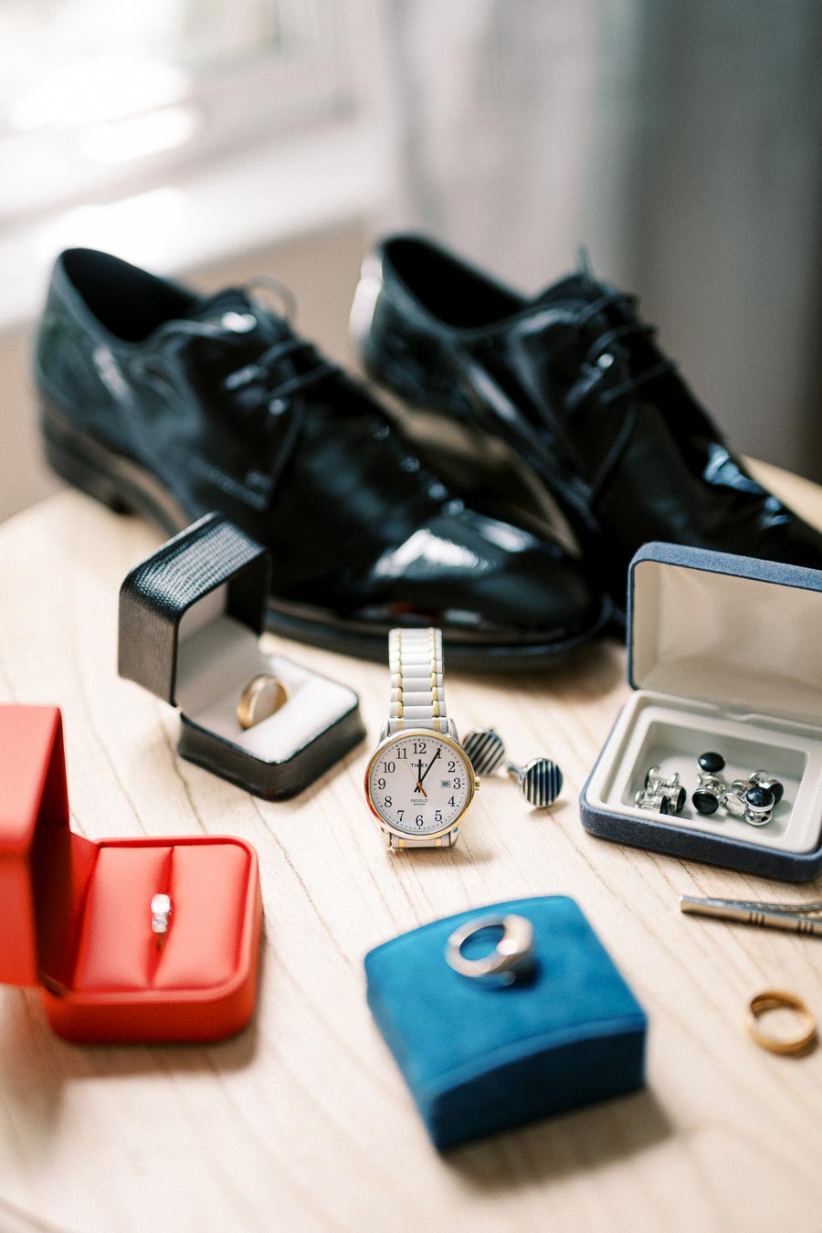 Elegant men's accessories arranged on a wooden table, featuring polished black dress shoes, a classic gold wristwatch, cufflinks, and several jewelry boxes containing rings, perfect for a groom's wedding day ensemble.