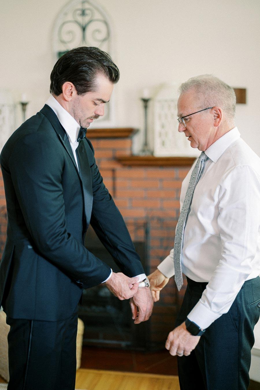 Two men getting ready for a formal event, one in a black suit adjusting his watch with the help of the other in a white shirt and tie, standing in a well-decorated room.
