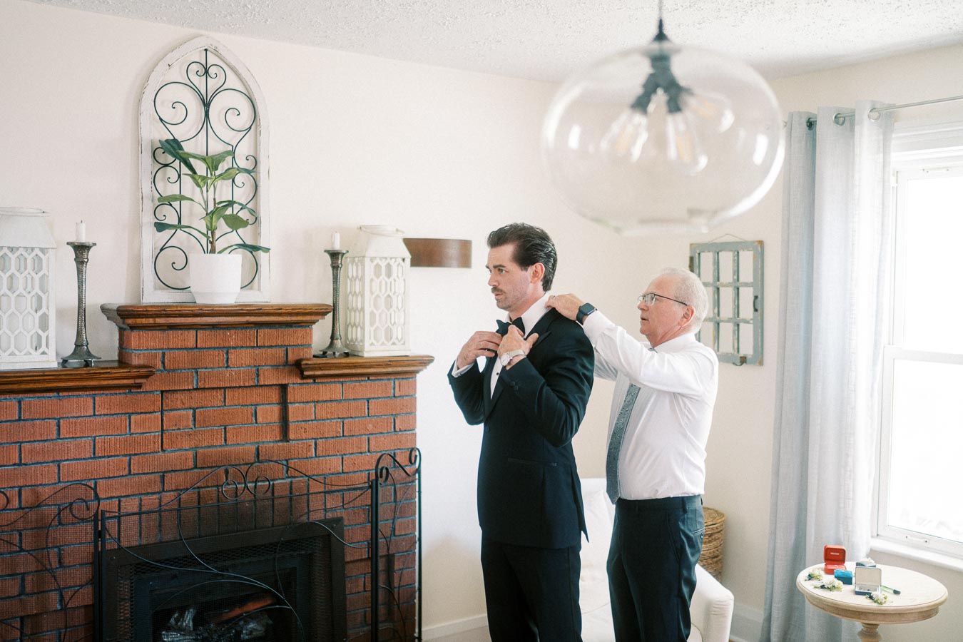 A man in a suit being helped with his tie by an older gentleman in a living room with a brick fireplace and decorative items.
