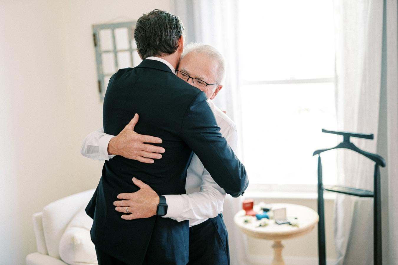 Father and son sharing an emotional embrace on wedding day, with formal attire, in a warmly lit room.