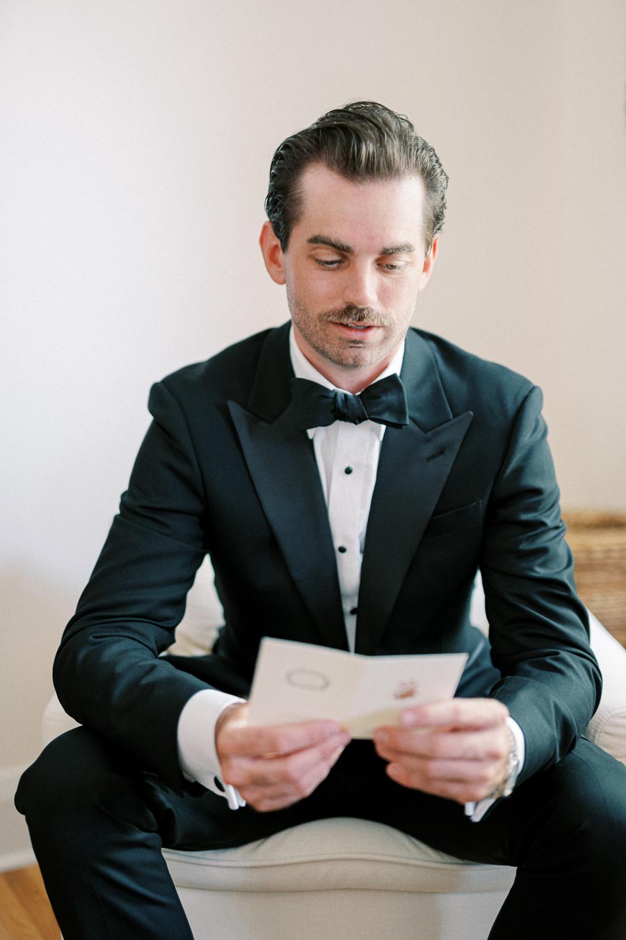 A man in a tuxedo sits in a chair, attentively reading a card, displaying a look of contemplation.