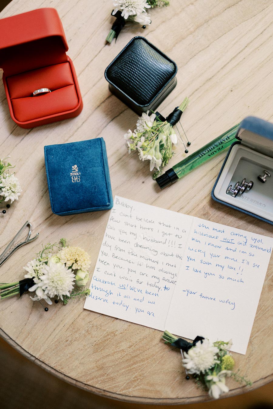 Wedding preparation flat lay with ring boxes, a handwritten note, white floral boutonnieres, cufflinks, and a pen on a wooden table.
