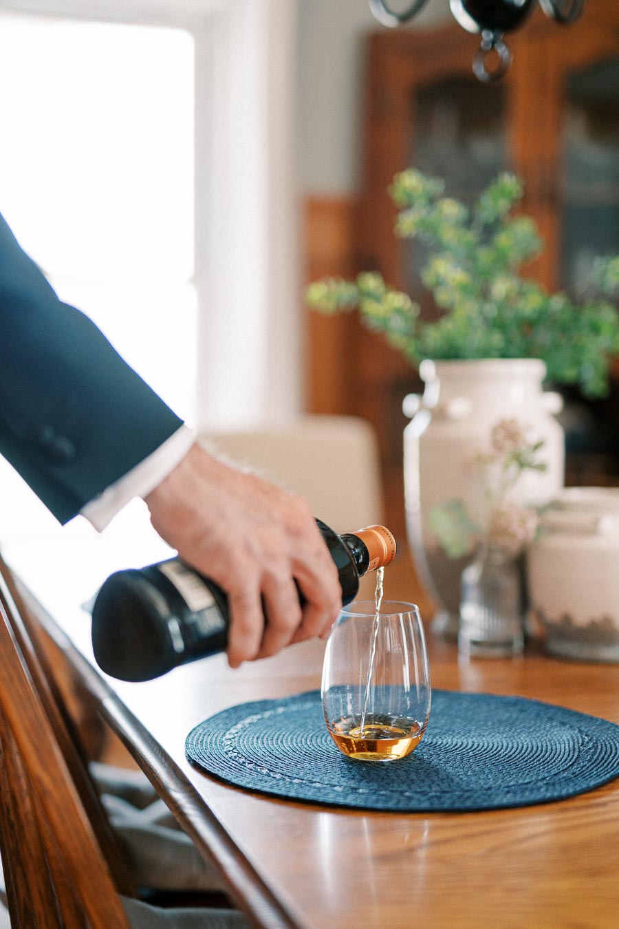 A person pours whiskey from a bottle into a tumbler glass on a wooden dining table with a blue placemat and decorative vases in the background.