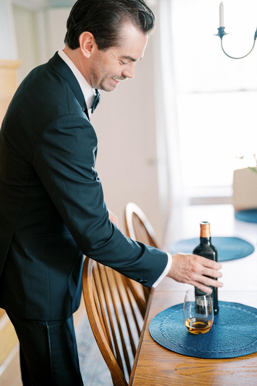 Man in a black suit placing a wine bottle and glass on a wooden dining table with blue placemats in a bright dining room.
