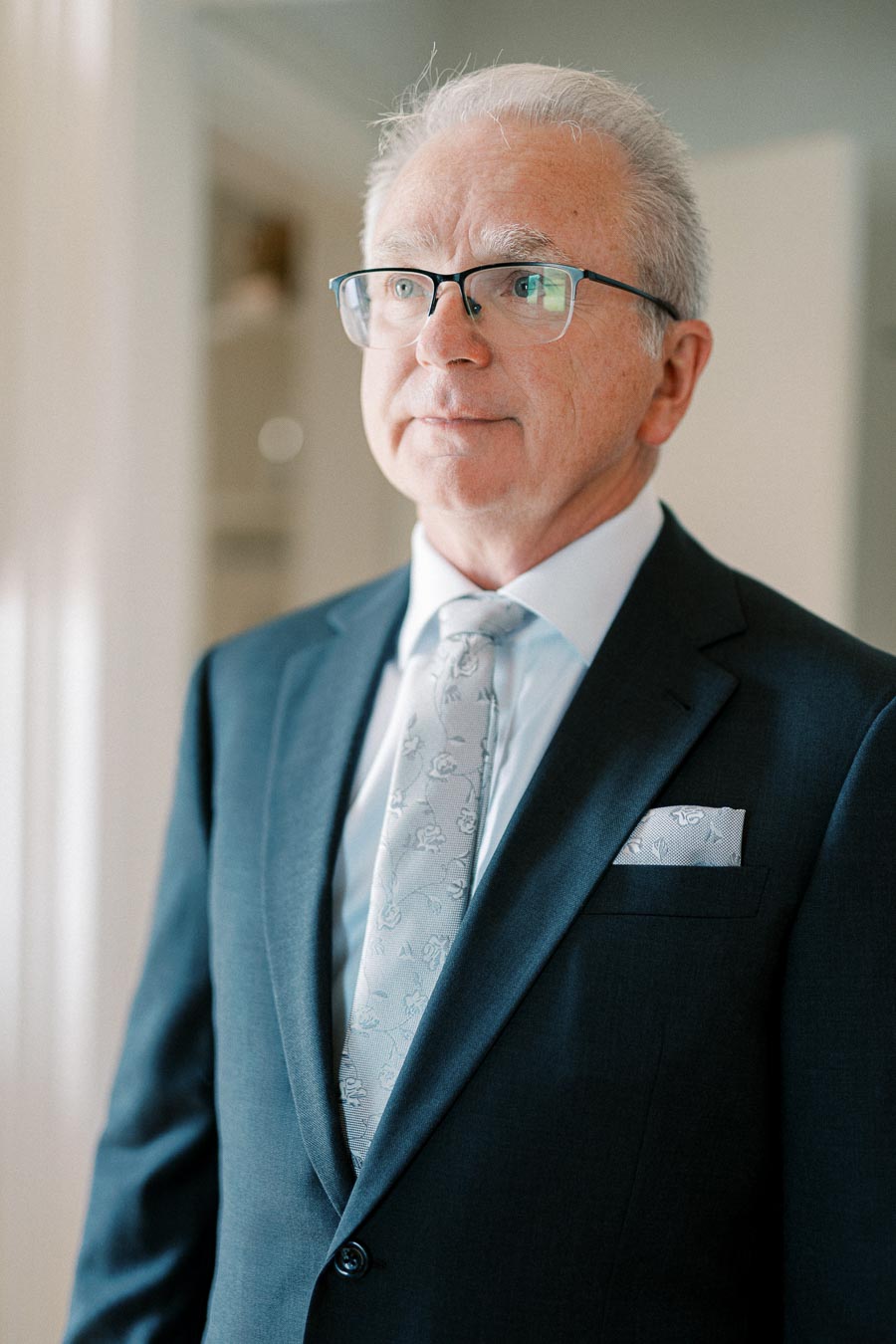 A distinguished elderly man wearing a suit, glasses, and a patterned tie standing indoors.