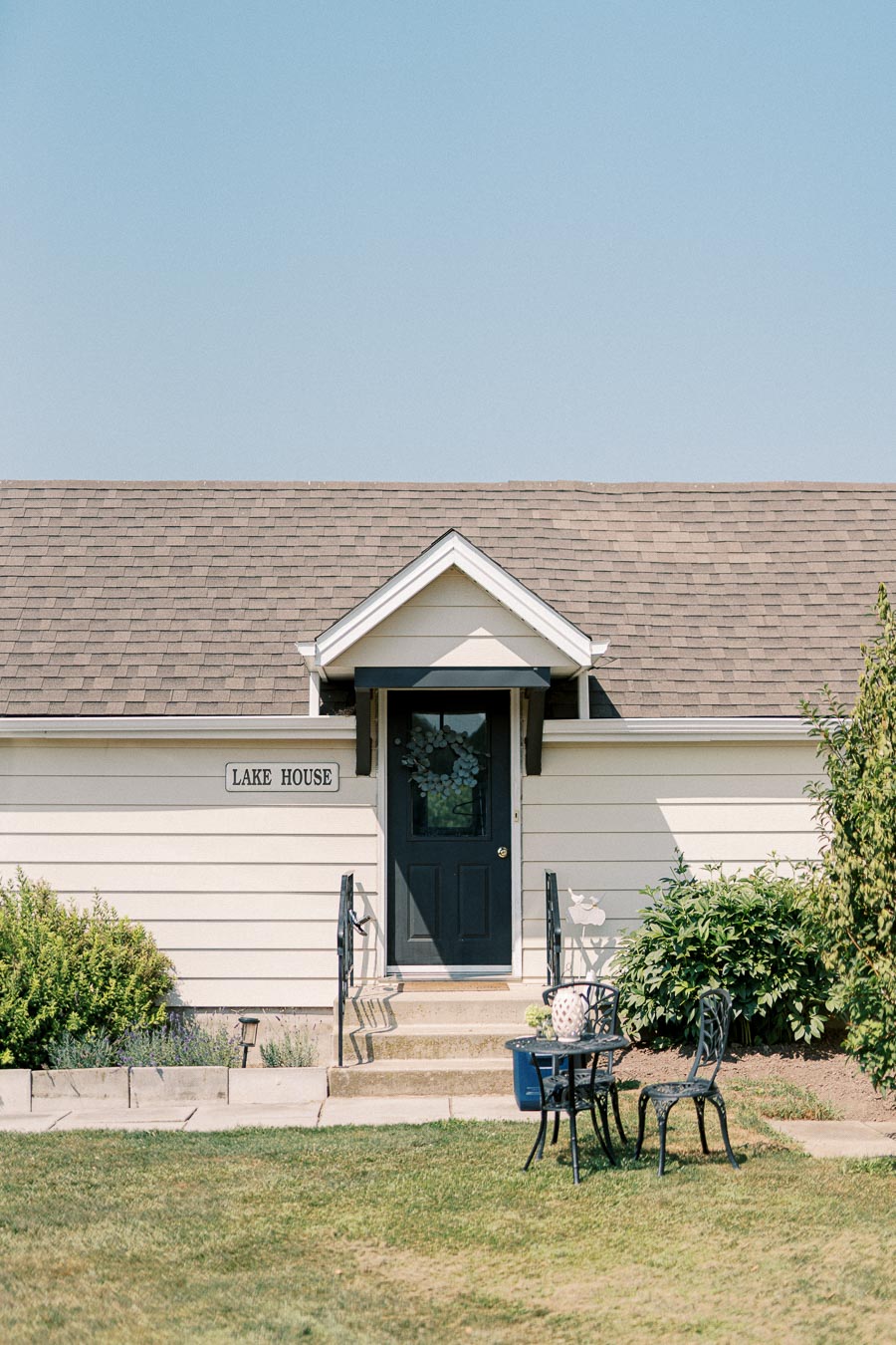 Front view of a charming light beige lake house with a dark door, surrounded by greenery and featuring an outdoor patio set, under a clear blue sky.